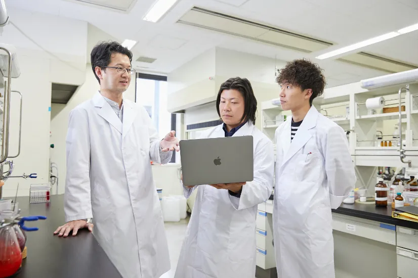 写真:さまざまな機材がある研究室内で、全員白衣姿の男性教員とノートパソコンを持った男子学生1名、説明を聞いている男子学生1名