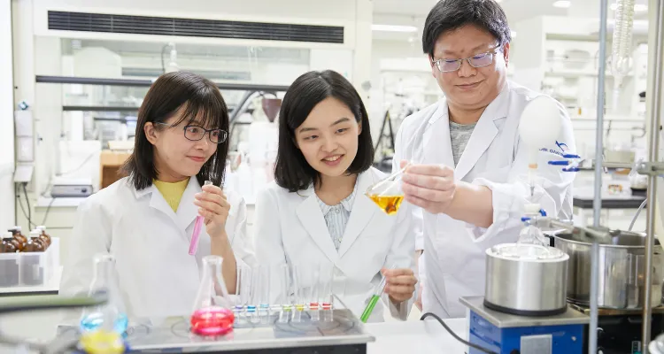 写真:白衣姿の生徒2名と教員がフラスコなどを手に取って研究している